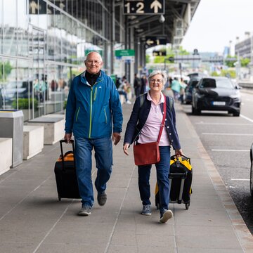 Zwei Personen mit Gepäck gehen entlang des Terminalgebäudes am Flughafen Stuttgart. 