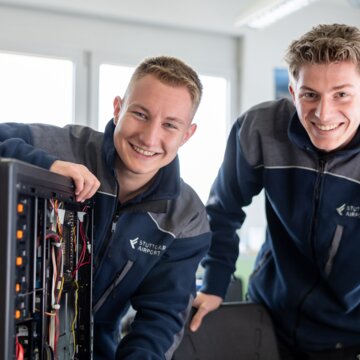 Zwei junge Männer in dunkelblauer Arbeitskleidung mit der Aufschrift "Stuttgart Airport" stehen in der Ausbildungswerkstatt hinter einem Tisch und lächeln in die Kamera. Vor ihnen steht ein geöffnetes Computergehäuse, über das sich eine der Personen leicht vorbeugt, während die andere sich über einen Stuhl lehnt. Auf dem Tisch liegen weitere technische Geräte und Unterlagen.