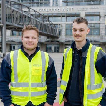 Zwei junge Männer in dunkelblauer Arbeitskleidung und neongelben Warnwesten stehen auf dem Vorfeld des Flughafens Stuttgart und haben die Hände in die Hüfte gestützt. Hinter ihnen befinden sich eine gläserne Fluggastbrücke, ein gelbes Vorfeldschild mit der Nummer 11 sowie das Terminalgebäude mit der Aufschrift ‚Stuttgart, Manfred Rommel Airport‘. 