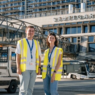 Zwei junge Mitarbeitende in neongelben Warnwesten stehen auf dem Vorfeld des Flughafens Stuttgart. Beide tragen blaue Jeans, weiße T‑Shirts und ein blaues Lanyard um den Hals mit befestigtem Flughafenausweis. Hinter ihnen verläuft eine gläserne Fluggastbrücke. Im Hintergrund befinden sich mehrere Vorfeldfahrzeuge, darunter ein Bus für den Passagiertransport, sowie das Terminalgebäude mit der Aufschrift ‚Stuttgart, Manfred Rommel Airport‘ unter einem blauen Himmel.