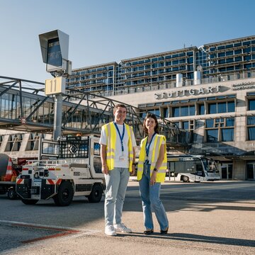 Zwei junge Mitarbeitende in neongelben Warnwesten stehen auf dem Vorfeld des Flughafens Stuttgart. Beide tragen blaue Jeans, weiße T‑Shirts und ein blaues Lanyard um den Hals mit befestigtem Flughafenausweis. Hinter ihnen verläuft eine gläserne Fluggastbrücke. Im Hintergrund befinden sich mehrere Vorfeldfahrzeuge, darunter ein Bus für den Passagiertransport, sowie das Terminalgebäude mit der Aufschrift ‚Stuttgart, Manfred Rommel Airport‘ unter einem blauen Himmel.