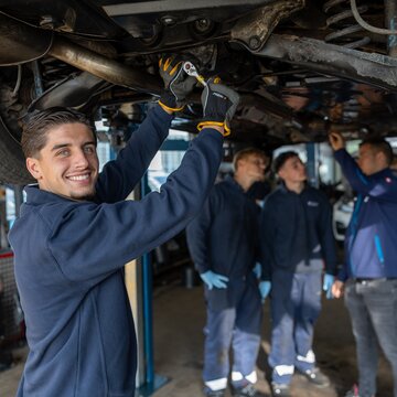 Ein junger Mitarbeitender arbeitet in einer Kfz‑Werkstatt an der Unterseite eines auf einer Hebebühne angehobenen Fahrzeugs und hält dabei ein Werkzeug. Im Hintergrund stehen drei weitere Personen in dunkelblauer Arbeitskleidung, die mit einer Taschenlampe die Unterseite des Autos begutachten.
