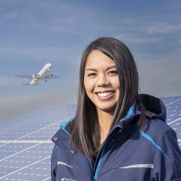 Eine junge Frau mit dunklen Haaren steht lächelnd vor einer Solaranlage auf dem Flughafengelände Stuttgart. Sie trägt eine dunkelblaue Jacke mit dem Logo „Stuttgart Airport“. Im Hintergrund hebt ein Flugzeug in den Himmel ab.