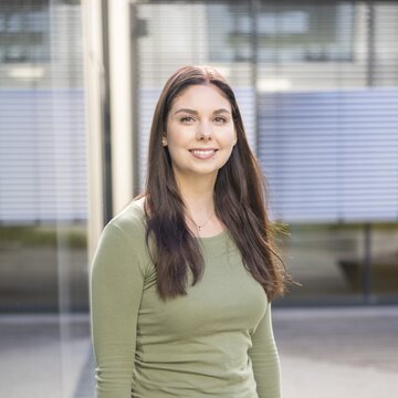 unge Frau steht lächelnd vor einem Bürogebäude mit Glasfassade am Flughafen Stuttgart.