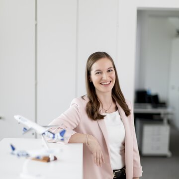 Junge Frau im Business-Outfit lächelt in einem modernen Büro neben zwei Modellflugzeugen.