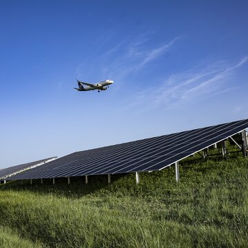 Bei strahlend blauem Himmel fliegt ein Flugzeug im Landeanflug über eine Solaranlage, die im Grünen steht.