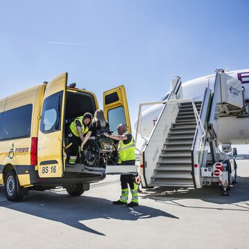Flughafenmitarbeiter helfen einer Person im Rollstuhl beim Einsteigen in ein Fahrzeug. 