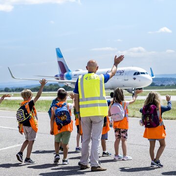 Kinder winken einem Flugzeug auf dem Flughafen Stuttgart Kinder in orangefarbenen Sicherheitswesten winken einem Flugzeug auf dem Flughafen Stuttgart zu.