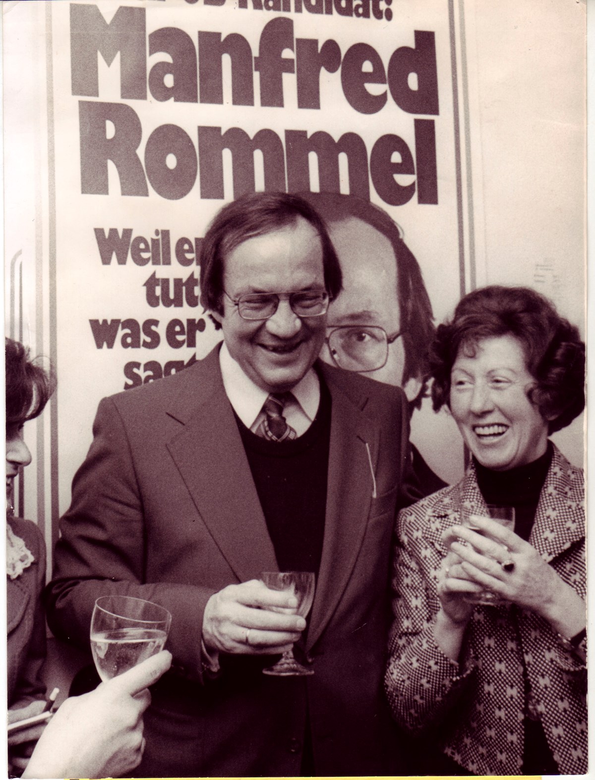 Manfred Rommel lacht gemeinsam mit einer Gruppe von Leuten, alle haben ein Sektglas in der Hand