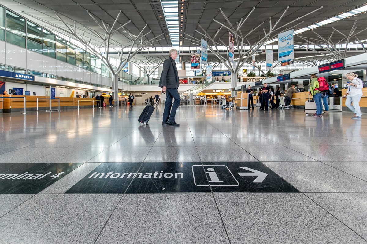 Mann mit Koffer läuft durch Terminal, im Hintergrund viele Menschen sowie Check-in-Schalter