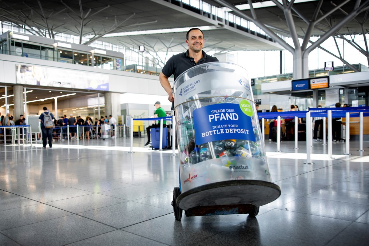 Ein Mann schiebt auf einem Wagen einen Sammelbehälter voller Flaschen durchs Terminal am Flughafen Stuttgart. Auf der Tonne steht „Spende dein Pfand – Donate your bottle deposite“.